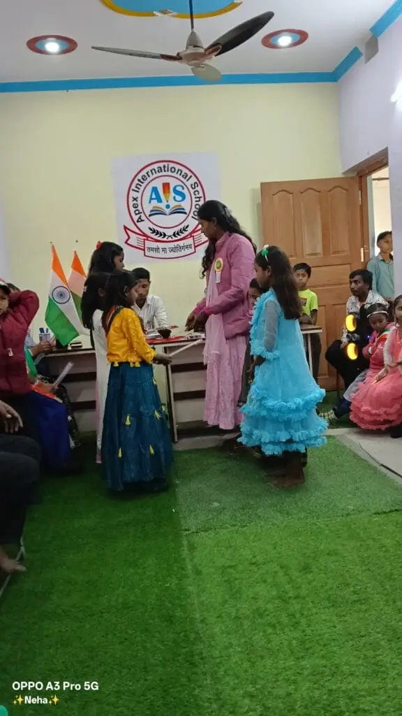 Best school in manigachhi, Indrathair and Darbhanga: An indoor student awards or prize distribution ceremony at Apex International School. This top cbse school in Indrathair, Manigachhi & Darbhanga celebrates student achievements, cultural participation, and academic milestones during school events. The image captures a female coordinator in a pink outfit handing out recognition on a green grass carpet stage. Young students in festive dresses—including a yellow and blue lehenga and a ruffled blue gown—patiently wait their turn.The official "AIS" Apex International School crest banner stands behind the main table alongside the Indian national flag. As a premier top school in Indrathair, Manigachhi & Darbhanga, the campus fosters confidence, performance, and leadership. This focus on student acknowledgment makes it the best english school in manigachhi, Indrathair and Darbhanga for a well-rounded education provided by the top english school in Indrathair, Manigachhi & Darbhanga.